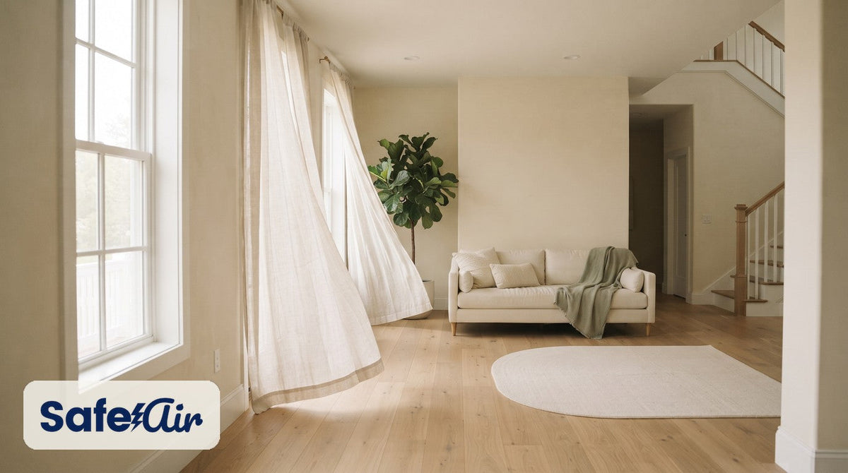 Sunlit cream living room with tall windows, sheer curtains, a sofa, and an oak staircase.
