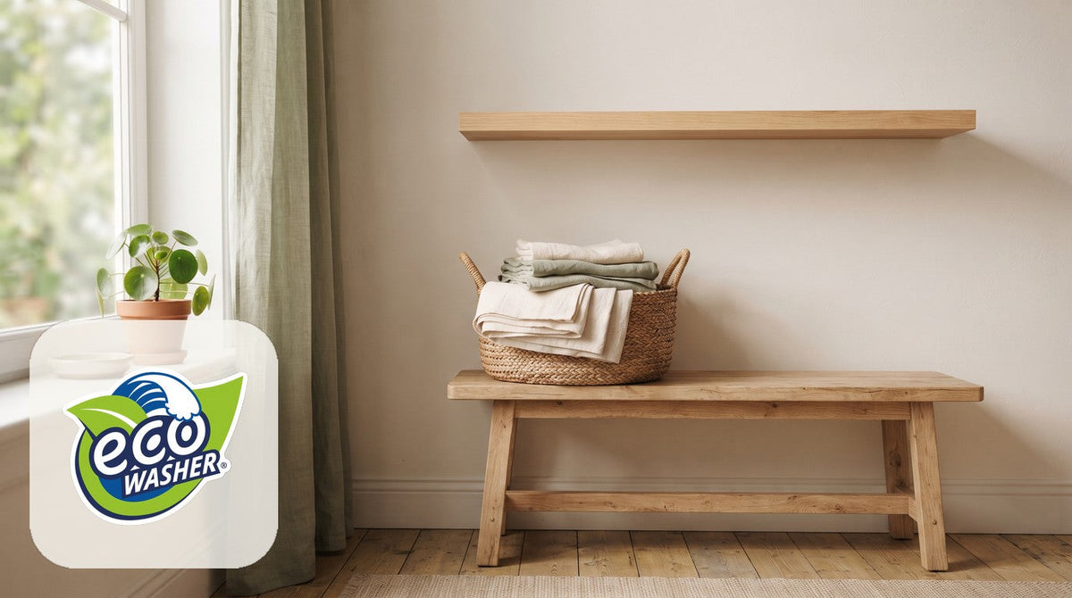 Calm laundry nook with a woven basket of folded cream and sage linens on a light wood bench beside a sunlit window