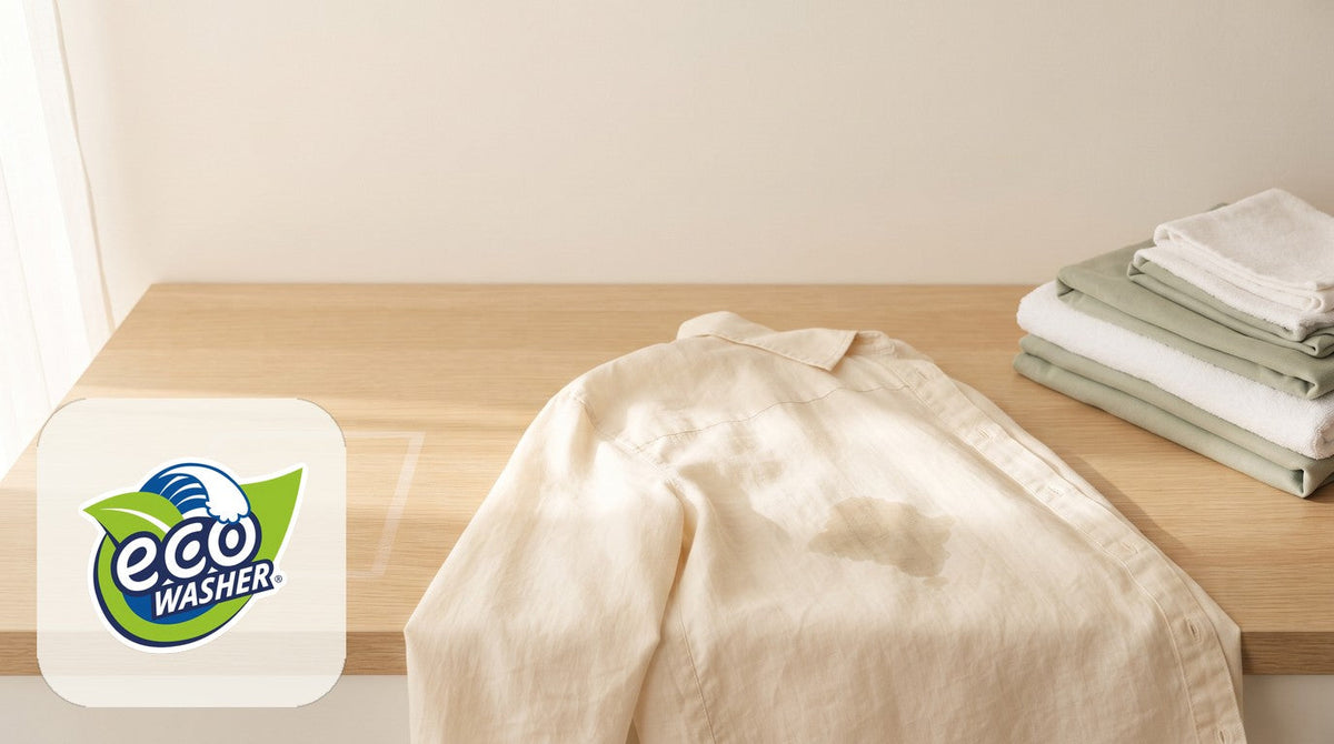 Cream linen shirt laid flat on a light oak counter with a damp spot, beside folded white and sage cloths.